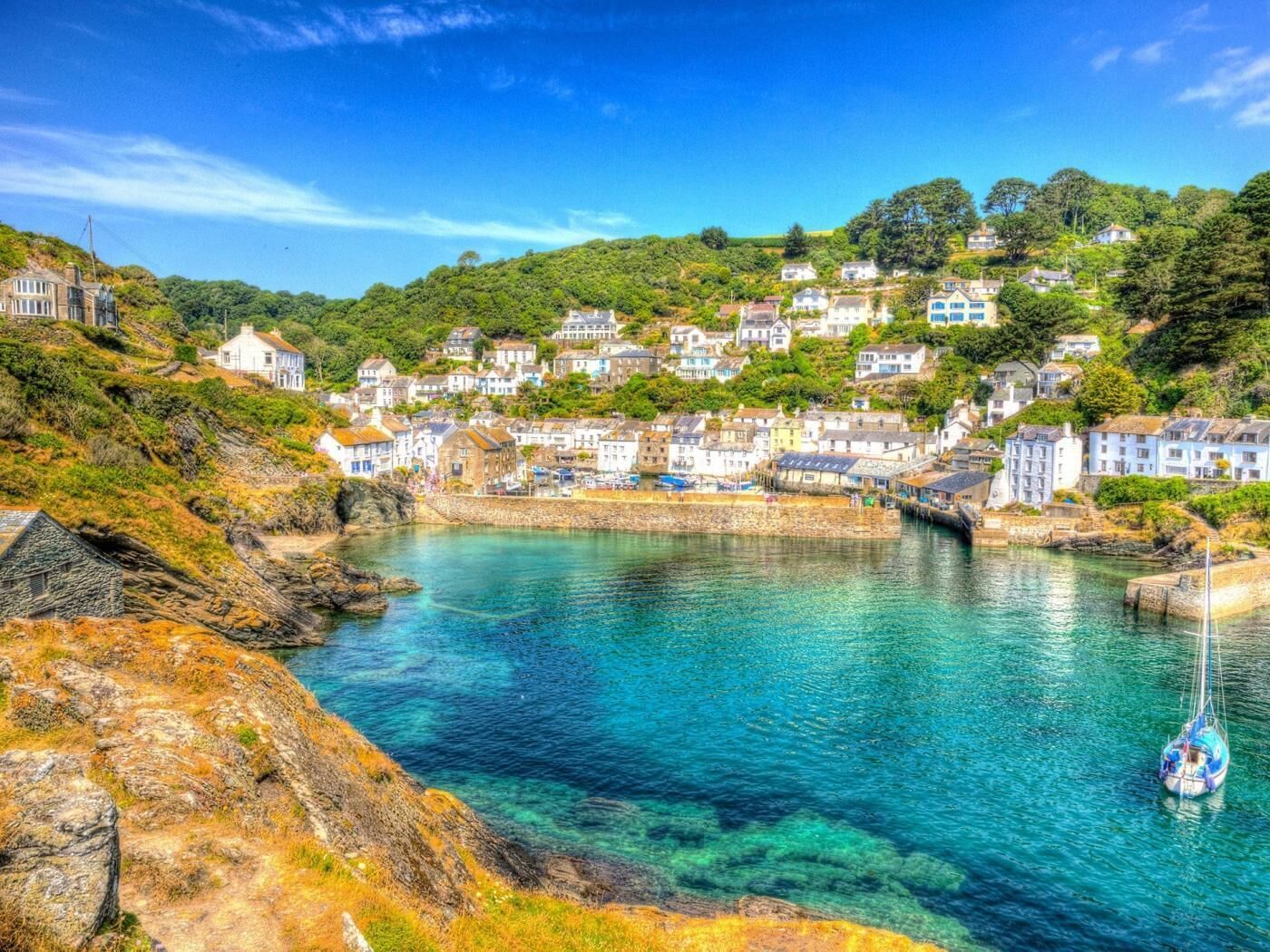A coastal village with houses on a hillside, a clear blue harbor, and a sailboat anchored near the shore.