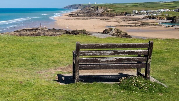 A bench on a grassy area overlooking a beach with a clear view of the ocean and distant buildings. - 4 Bed in Holsworthy (oc-lamer) (Holsworthy Hamlets)