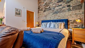 Una camera da letto con una parete in pietra, un letto con testiera blu, un comodino in legno e una poltrona in pelle.
