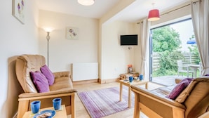 Ein modernes Wohnzimmer mit einer Couch, einem Sessel und einem Tisch mit Keksen. Ein Heizkörper, ein an der Wand befestigtes Fernsehgerät und ein Fenster mit Vorhängen sind ebenfalls vorhanden.