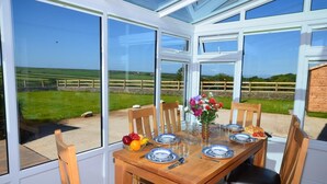 A conservatory with a table set for four, chairs, a vase with flowers, and a view of the countryside. - 3 Bed in Bideford (oc-glake) (Hartland)
