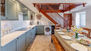 A modern kitchen with a built-in washer and dryer, a dining area with a wooden bench, and a staircase with wooden railings. - 5 Bed in Redruth (oc-hellf) (Illogan)