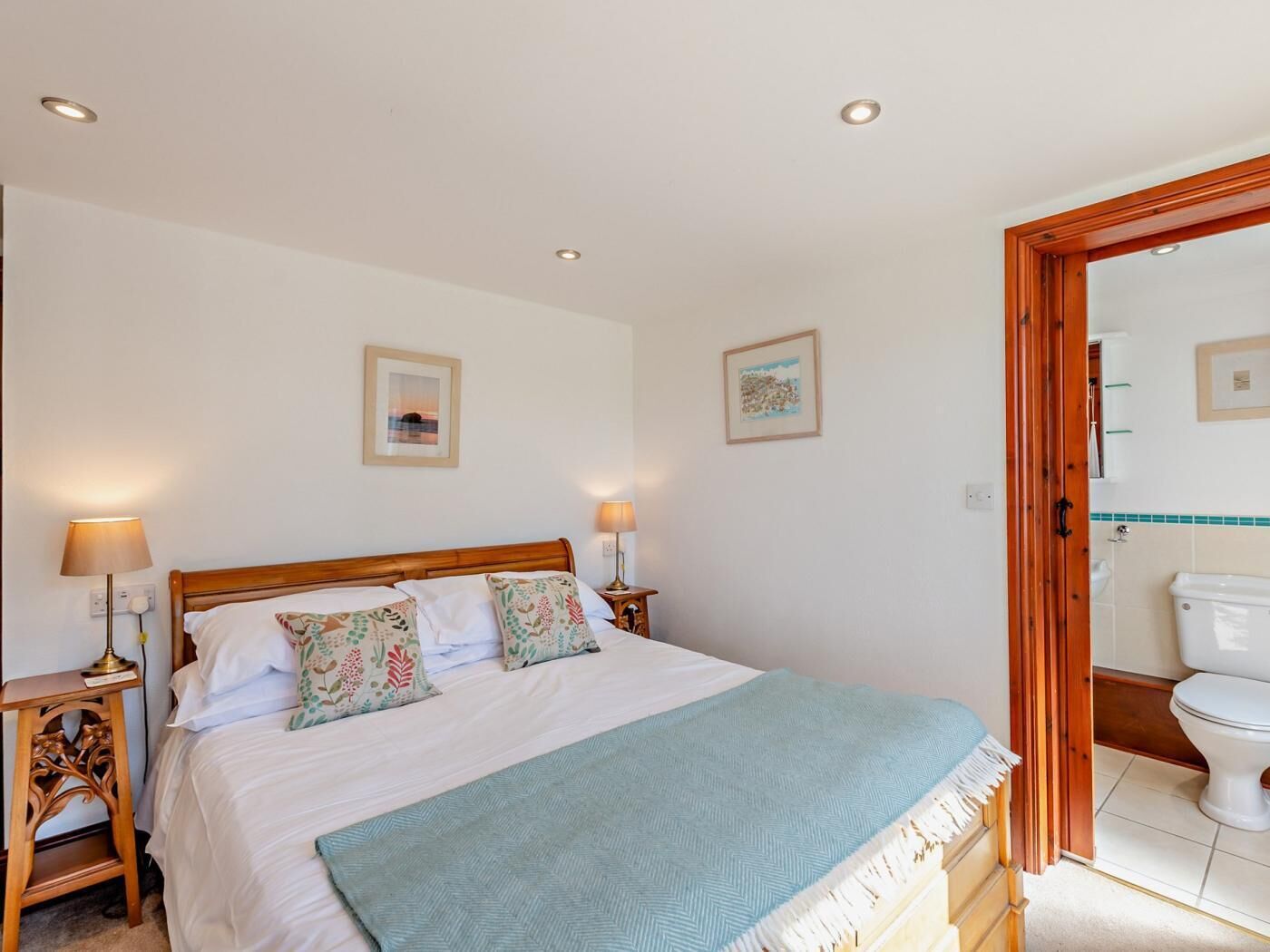 A bedroom with a wooden bed, two bedside tables with lamps, a blue throw blanket, and two framed pictures on the wall.