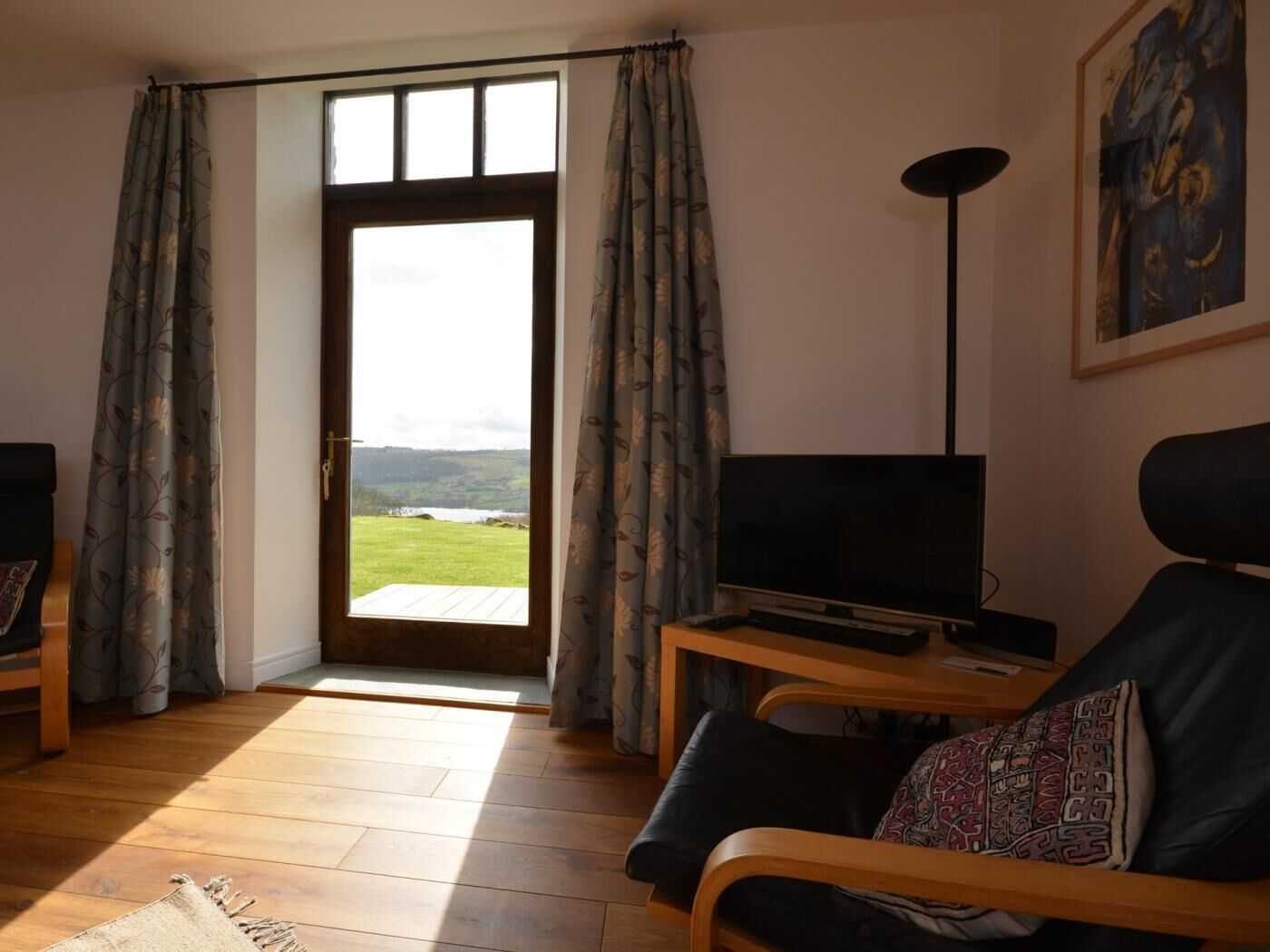 A living room with a TV, a sofa, a lamp, and a view of a landscape through a door.