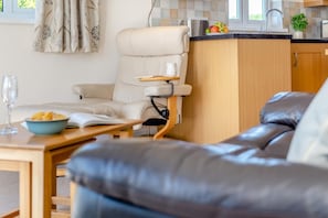 Ein modernes Wohnzimmer mit einer Ledersofa, einem Relaxsessel mit Tablett und einem Esstisch mit einer Schüssel Snacks, sowie eine Küchenzeile mit einer Obstschale.