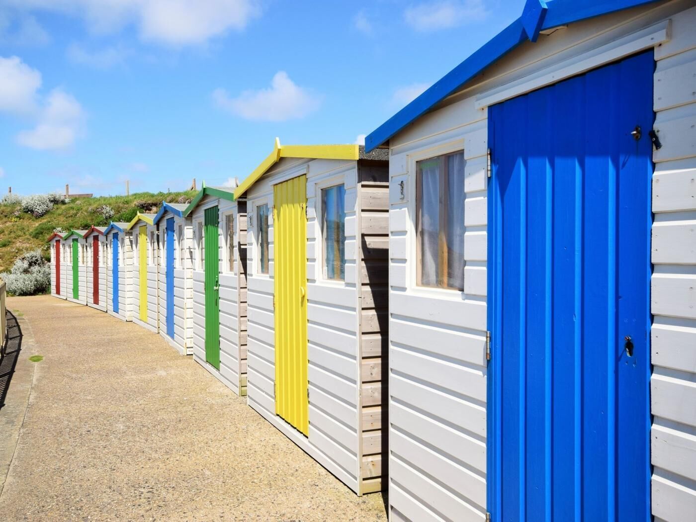 Eine Reihe bunter Strandhäuser mit weißer Verkleidung und blauen Türen, die an einem Kiesweg aufgereiht sind.