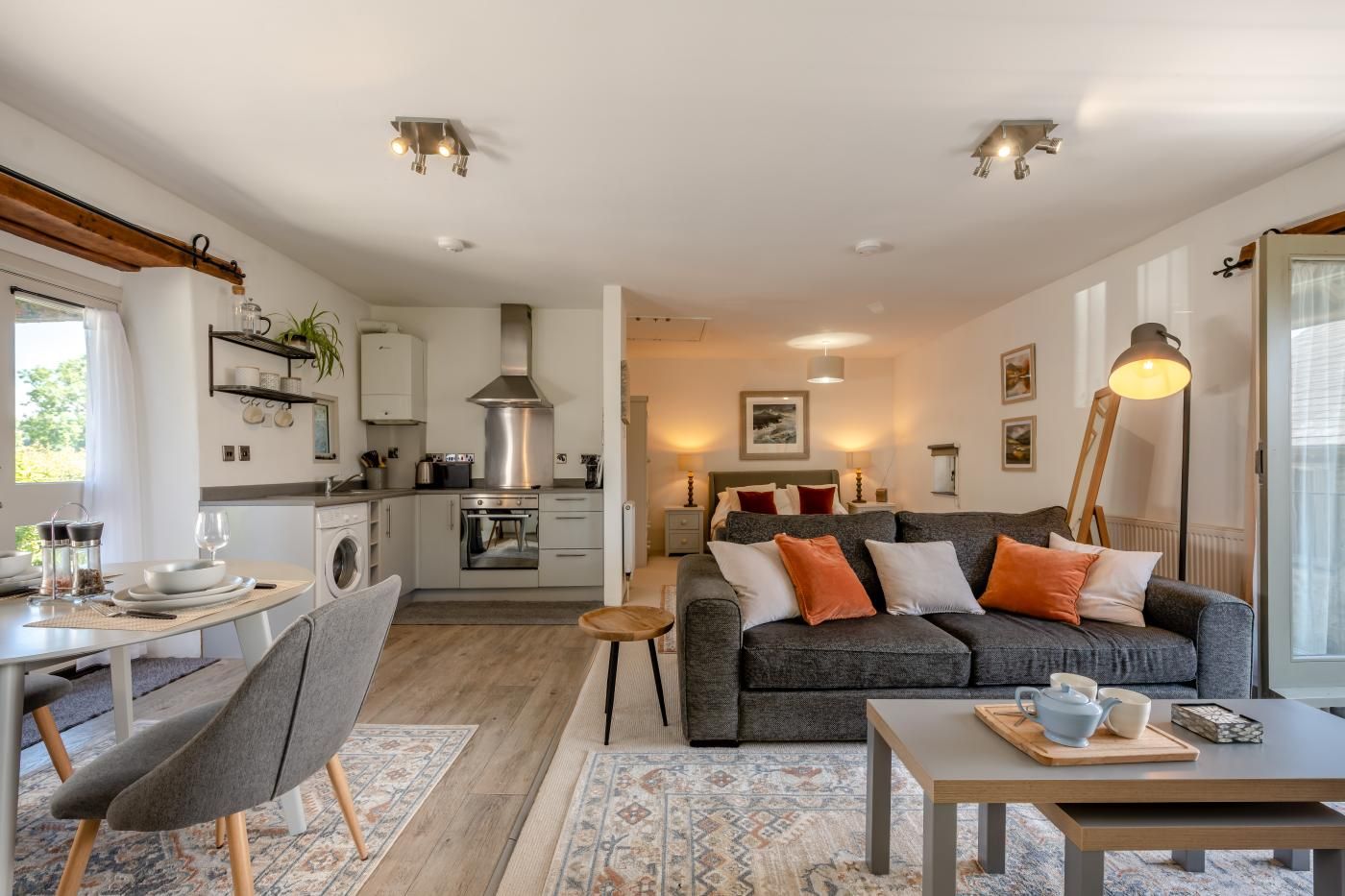 A modern living room with a grey sofa, a dining area with a table and chairs, and a kitchen with white cabinets and stainless steel appliances.