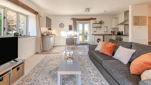 A modern living room with a sofa, coffee table, and TV, leading to a kitchen and dining area with a view of the outdoors. - 1 Bed in Tavistock (oc-highm) (Cudlipptown)