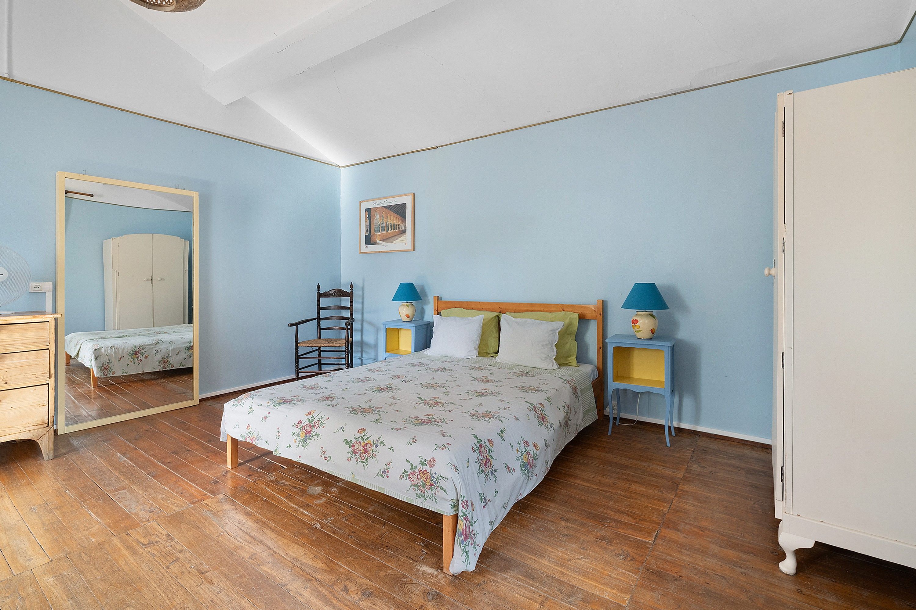 A bedroom with a wooden bed, a nightstand with a lamp, a dresser, a chair, and a mirror on the wall.