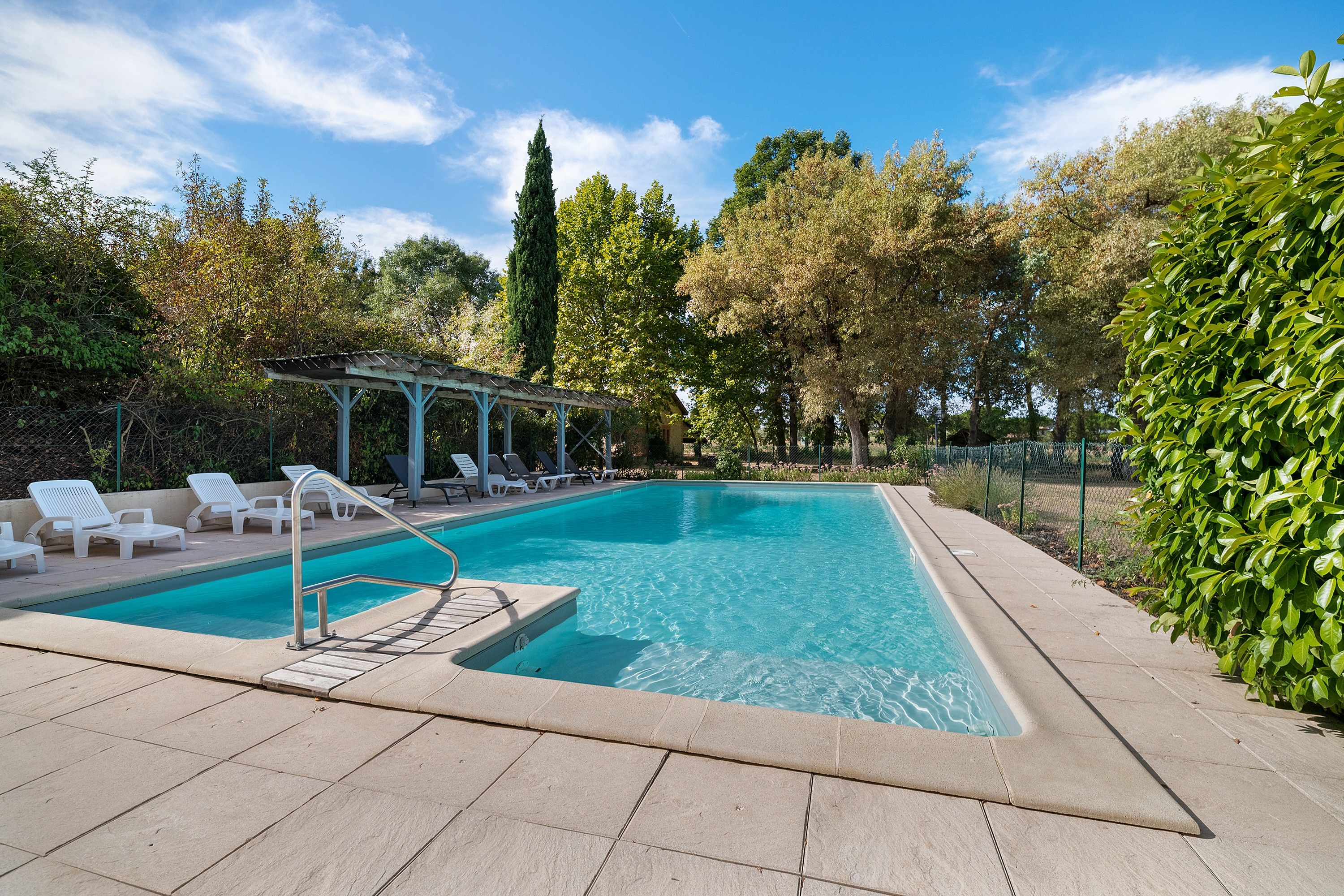A rectangular swimming pool with a tiled deck, surrounded by greenery and trees.