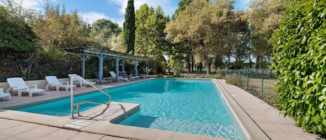 A rectangular swimming pool with a tiled deck, surrounded by greenery and trees.