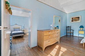 A room with a wooden dresser, a bed with a checkered bedspread, a chair, and a small table with a lamp.