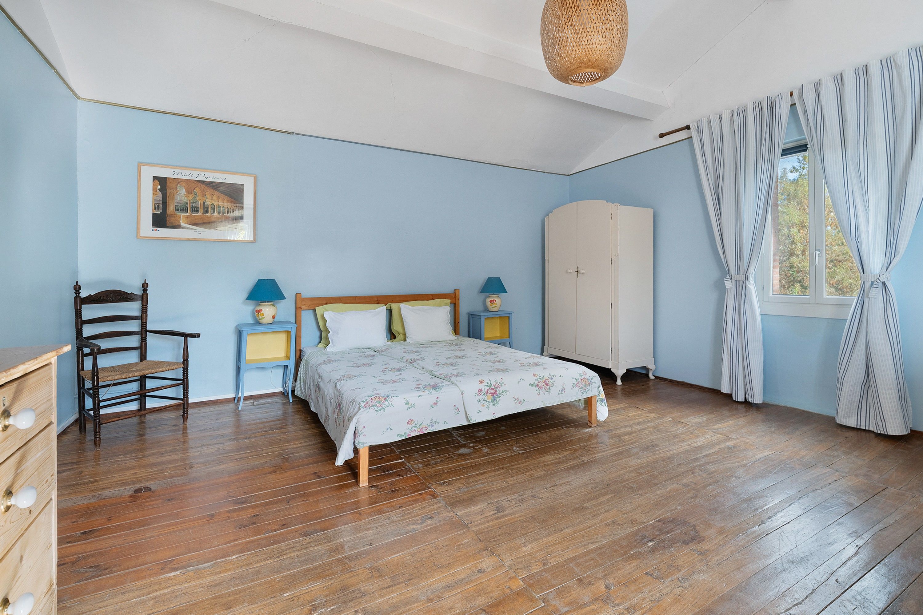 A bedroom with a wooden bed, a chair, a wardrobe, a bedside table with lamps, and a framed picture on the wall.