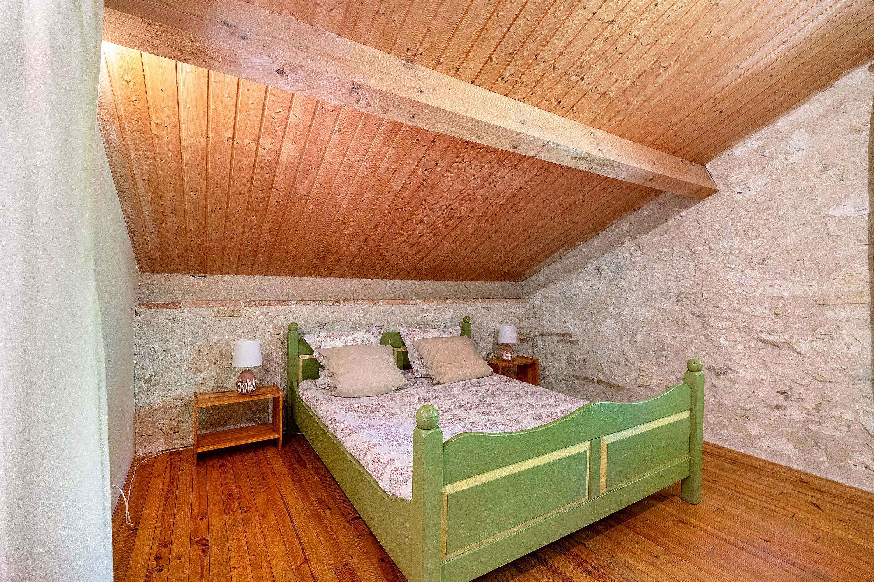 Ein Schlafzimmer mit schrägem Dach, Holzboden, einem grünen Bett und einer Steinwand.