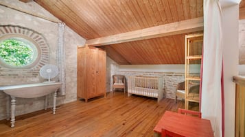 Una habitación rústica con techo de madera, un ventanal redondo, un lavamanos blanco, un armario de madera, una cuna y una mesa roja.