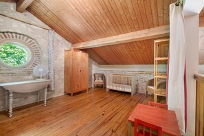 Une pièce rustique avec un plafond en bois, une fenêtre ronde, un lavabo blanc, une armoire en bois, un berceau et une table rouge.