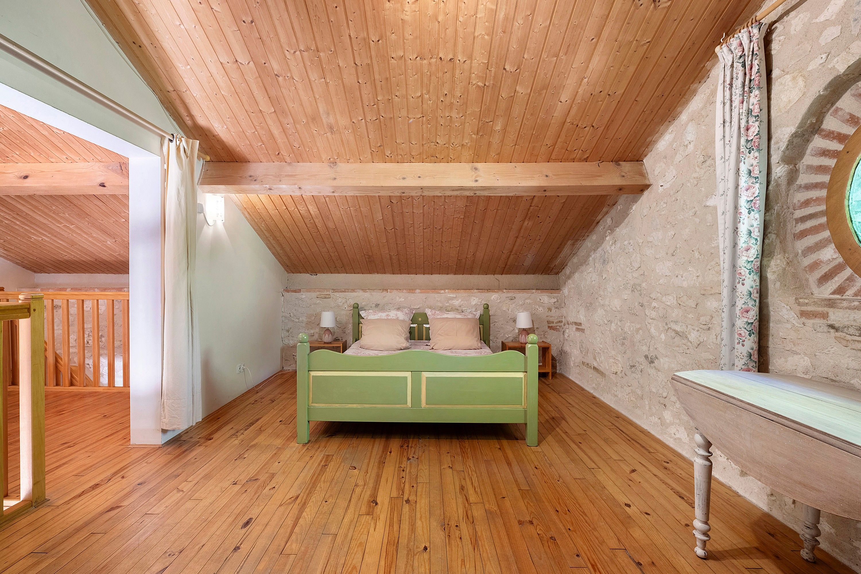 A bedroom with a wooden ceiling, a green bed, two bedside tables with lamps, and a stone wall.