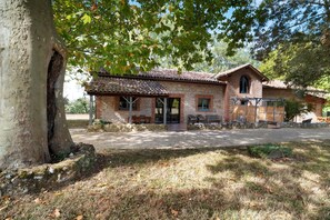 Un bâtiment rustique en pierre, doté d’un toit en tuiles, d’un porche avec des bancs et d’un grand arbre au premier plan.