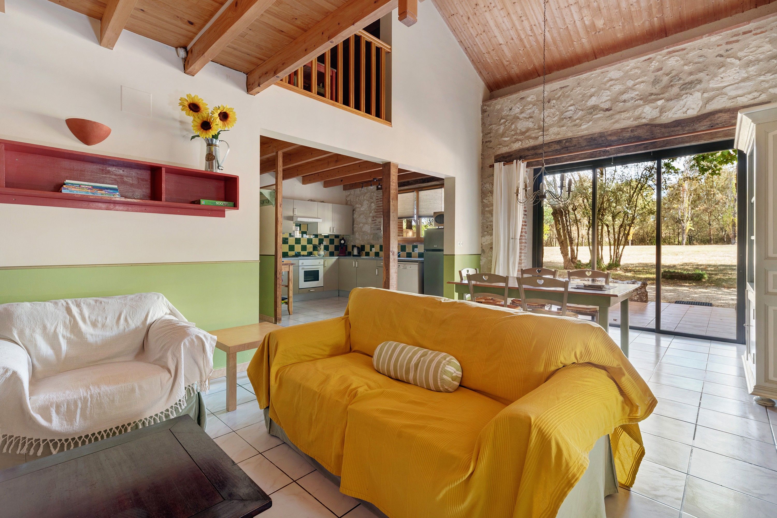 A spacious living area with a yellow sofa, a white armchair, and a small table. A kitchen is visible in the background, and there is a large window with a view of trees.