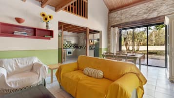 Amplia sala de estar con un sofá amarillo, un sillón blanco y una pequeña mesa. Se ve una cocina al fondo, y hay un ventanal con vistas a árboles.