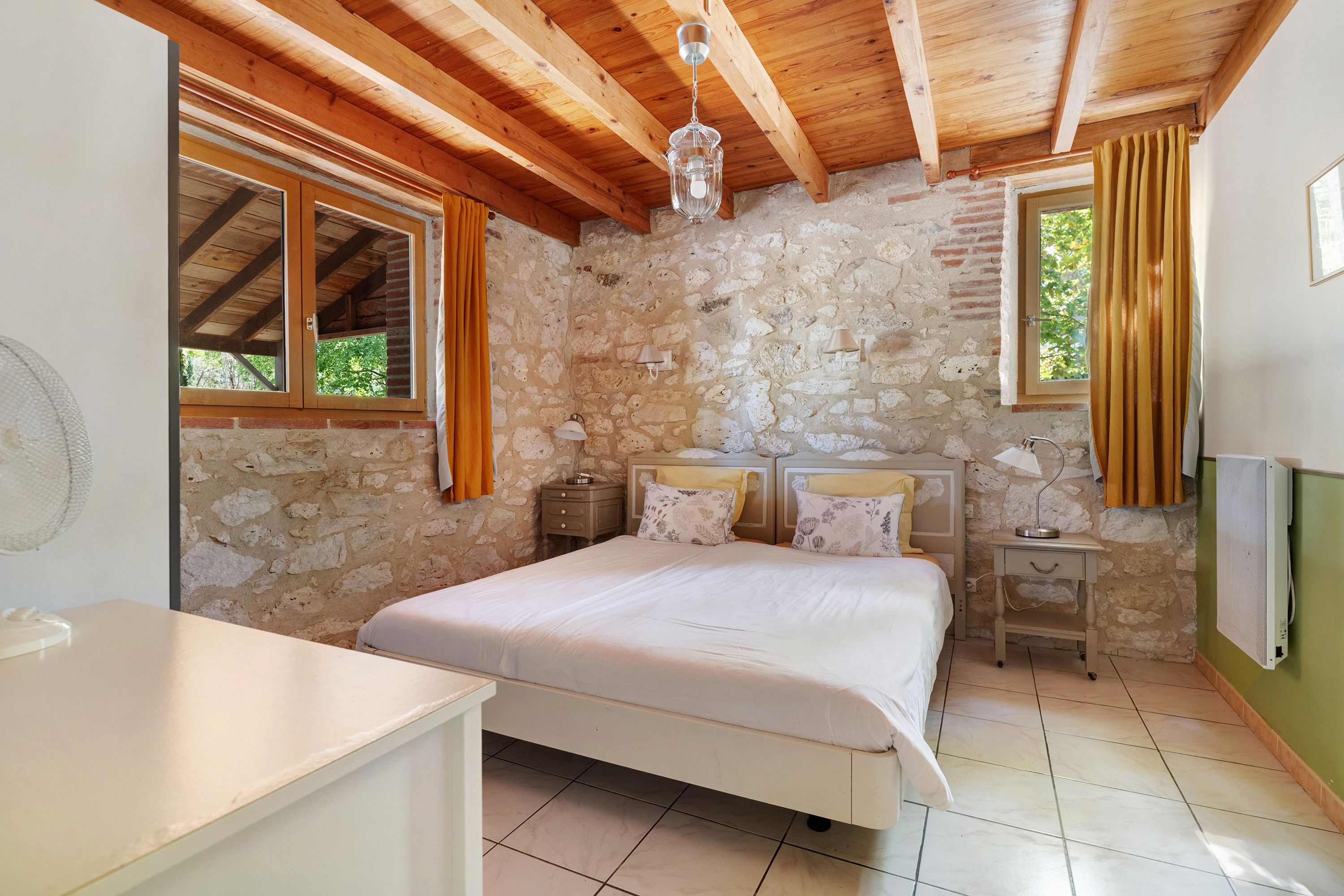 A bedroom with a stone wall, wooden ceiling beams, a bed with white bedding, two bedside tables with lamps, and a window with curtains.