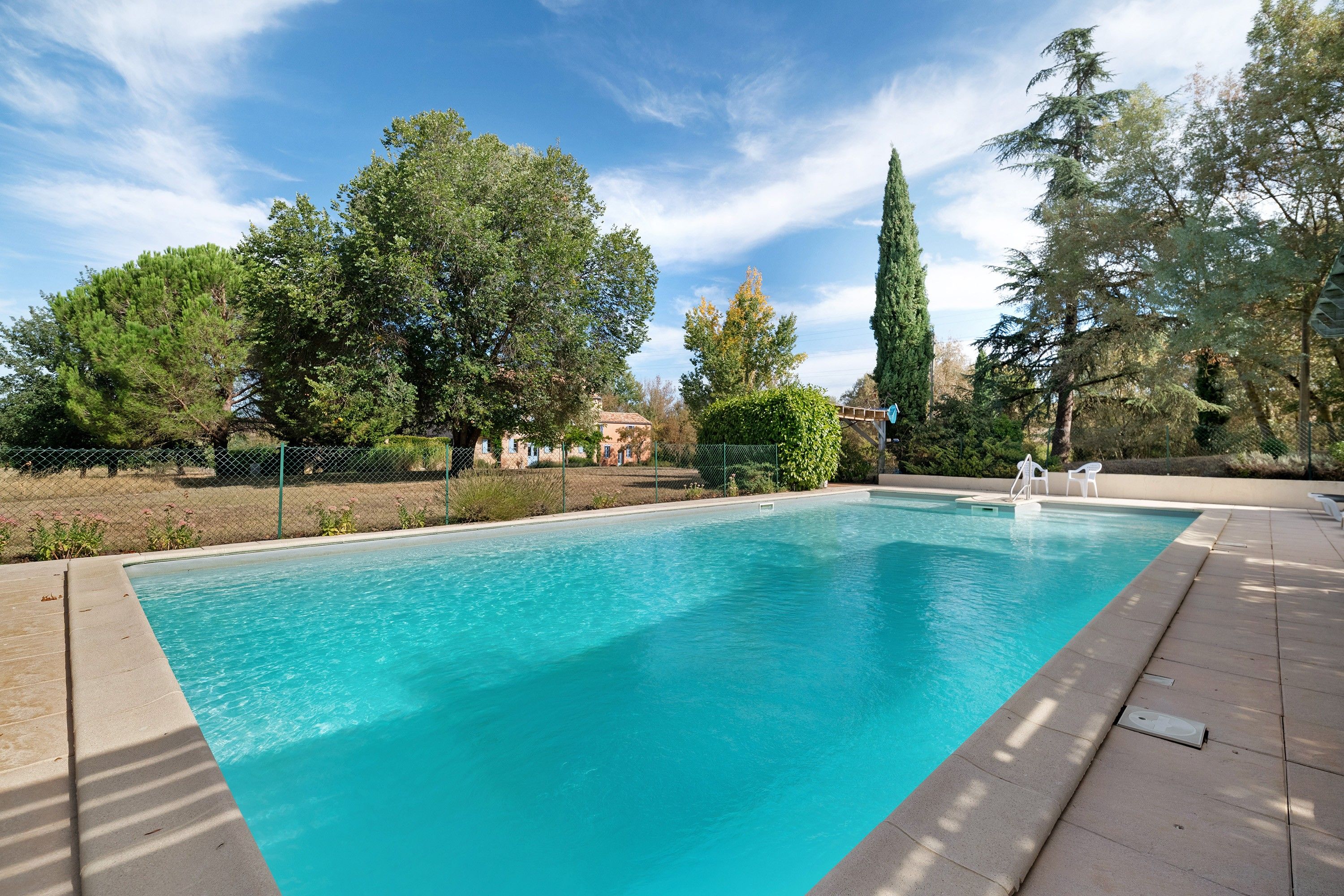 A rectangular swimming pool with clear blue water, surrounded by a fence and trees.