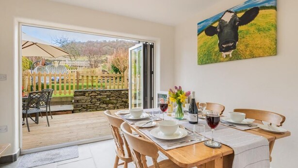 Zona pranzo con tavolo apparecchiato per quattro persone, un quadro di una mucca al muro e vista su un giardino attraverso una porta aperta.