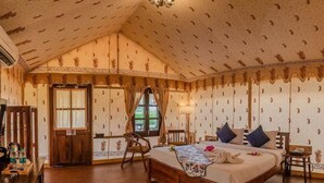 A bedroom with a wooden bed, a desk, and a chair. There is a window with curtains, a chandelier, and patterned wallpaper. - Desert Dhora Luxury Resort Jaisalmer (Jaisalmer)