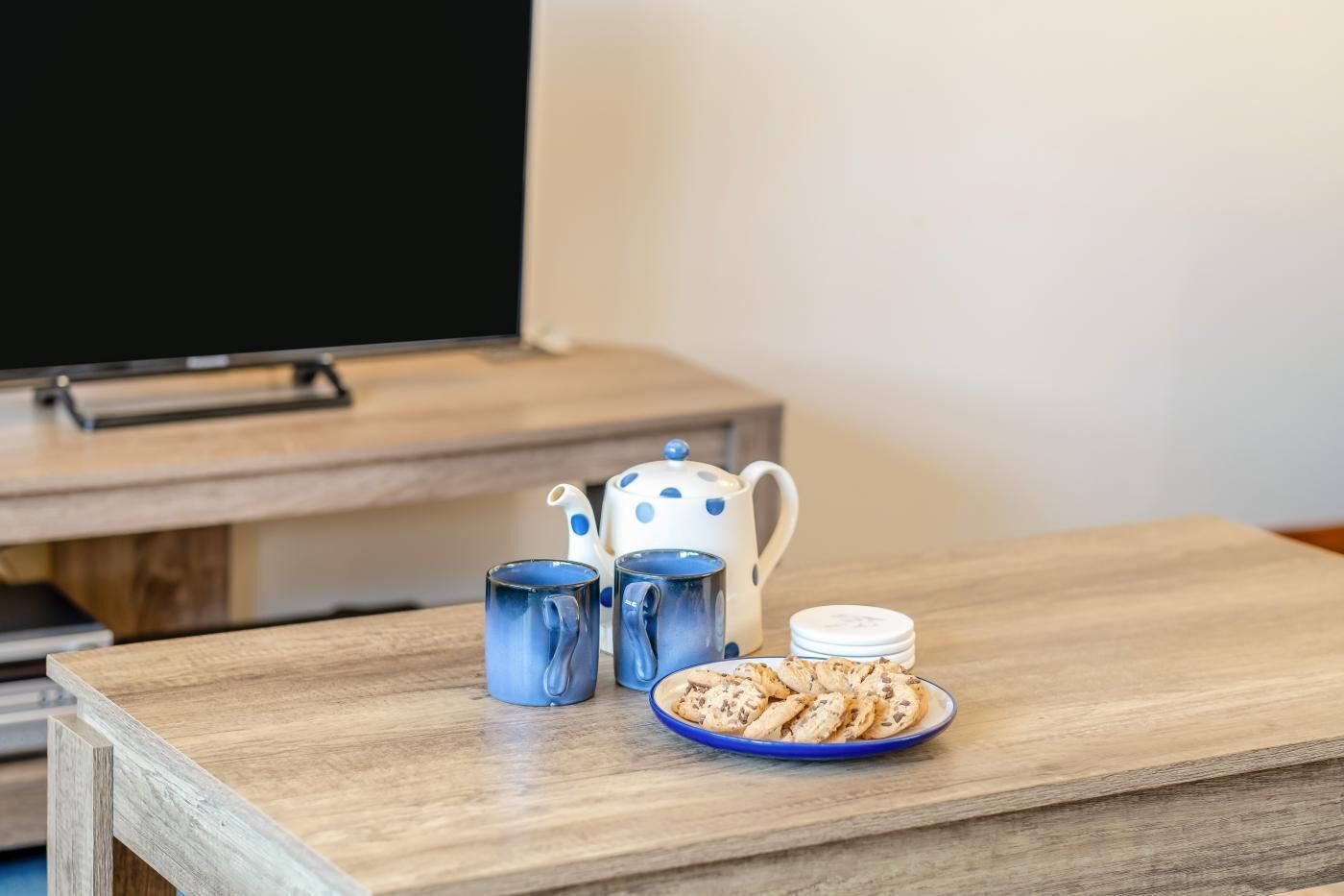 A wooden table with a plate of cookies and two blue mugs, a white teapot with blue polka dots, and a black screen in the background.