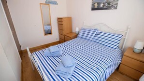 Ein Einzelbett mit einer blau-weißen gestreiften Bettdecke, zwei gefaltete Handtücher, ein Nachttisch mit Lampe, eine Holzkommode, ein Spiegel und ein an der Wand befestigtes Schild.