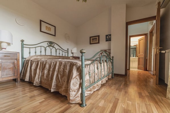 A bedroom with a wooden floor, a metal bed frame, a bedside table with a lamp, and a door leading to another room.
