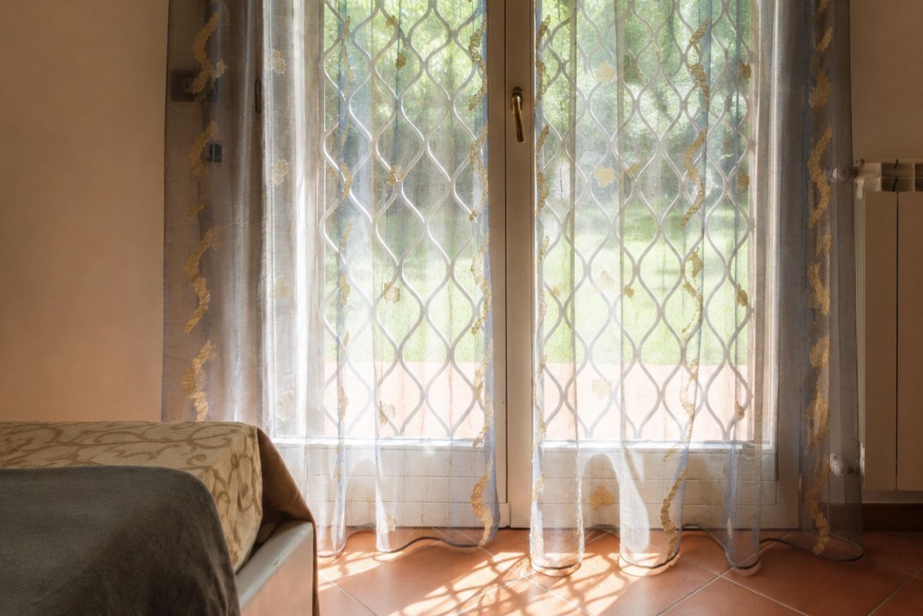 A room with a bed, a window with a patterned curtain, and a view of greenery outside.