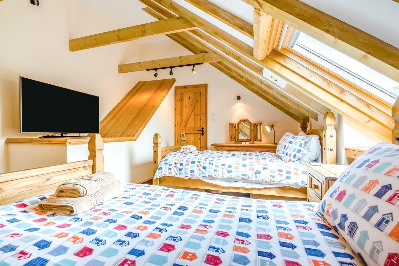 A bedroom with a sloped ceiling, wooden beams, a bed with patterned bedding, a bedside table, a TV, and a mirror.