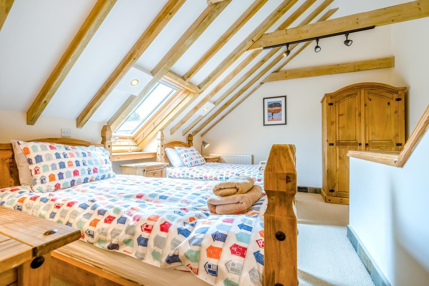 A bedroom with a sloped ceiling, wooden beams, a bed with patterned bedding, a bedside table, a wooden wardrobe, and a framed picture on the wall.