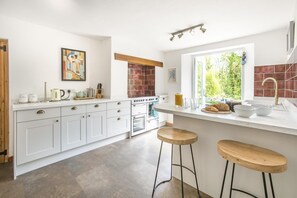 A modern kitchen with white cabinets, a large island with stools, a stove, and a window with a view of greenery. - 3 Bed in Bideford (oc-oldsb) (Buck's Mills)