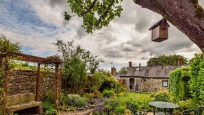 A stone pathway leading to a rustic wooden pergola, surrounded by lush greenery and a stone house with a sloped roof. - 2 Bed in Matlock (oc-pk885) (Winster)