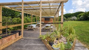 A wooden deck with picnic tables and planters, leading to a covered area with a building in the background. - 1 Bed in Par- (oc-sybar) (Saint Blazey)