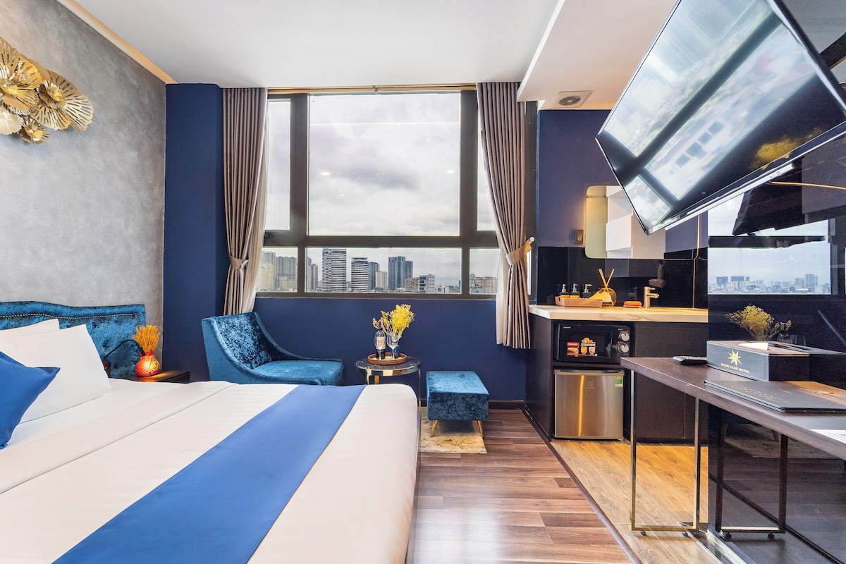 Ein Hotelzimmer mit einem großen Bett, einem Schreibtisch, einem Fernseher, Blick auf die Stadt und einer Küchenzeile.