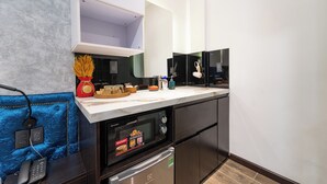 A compact kitchen with a microwave, oven, and built-in refrigerator. There is a phone on a blue upholstered chair, and a small table with a basket and a bottle. - Home Park Hotel HCM (Ho Chi Minh City)