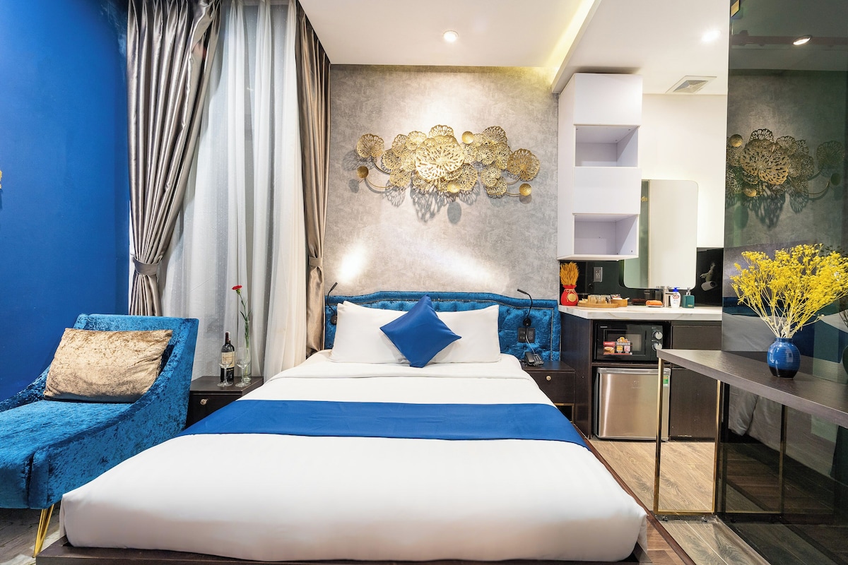 Ein modernes Hotelzimmer mit einem großen Bett, einem blauen Sessel, einer Küchenzeile und einem Wandbild.