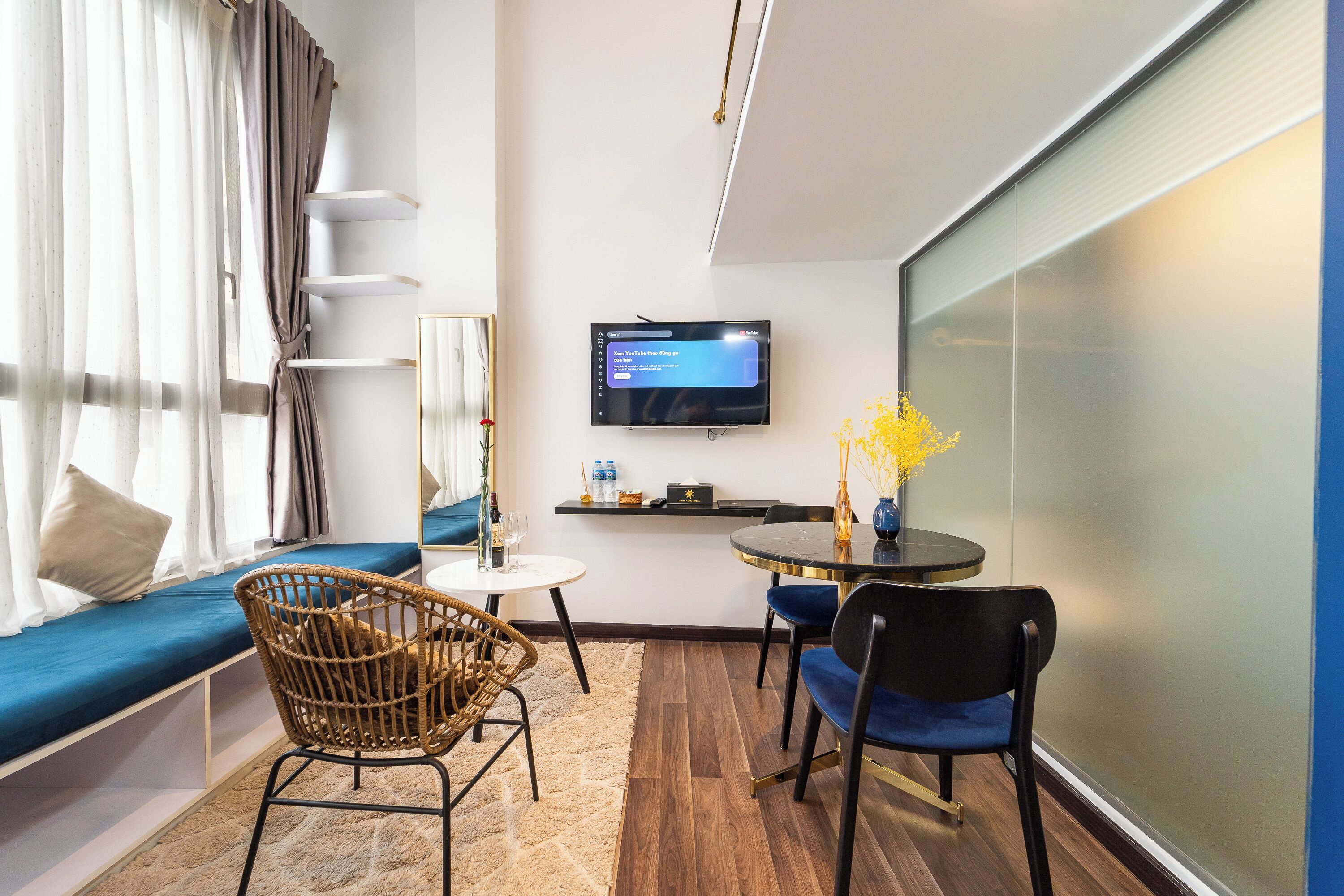 A modern hotel room with a sofa, two chairs, a small table, a TV, and a glass wall.