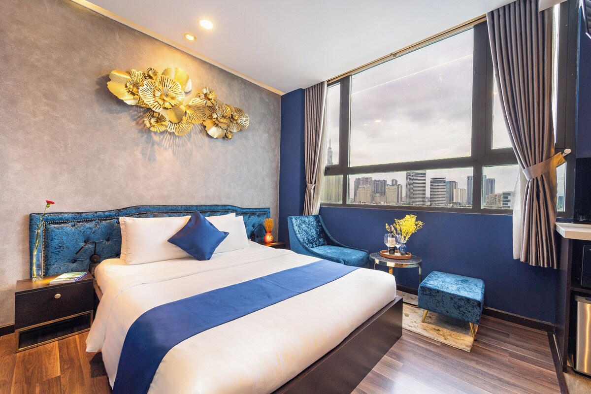 Een moderne hotelkamer met een groot bed, uitzicht op de stad, een blauwe accentmuur en een decoratief kunstwerk aan de wand.