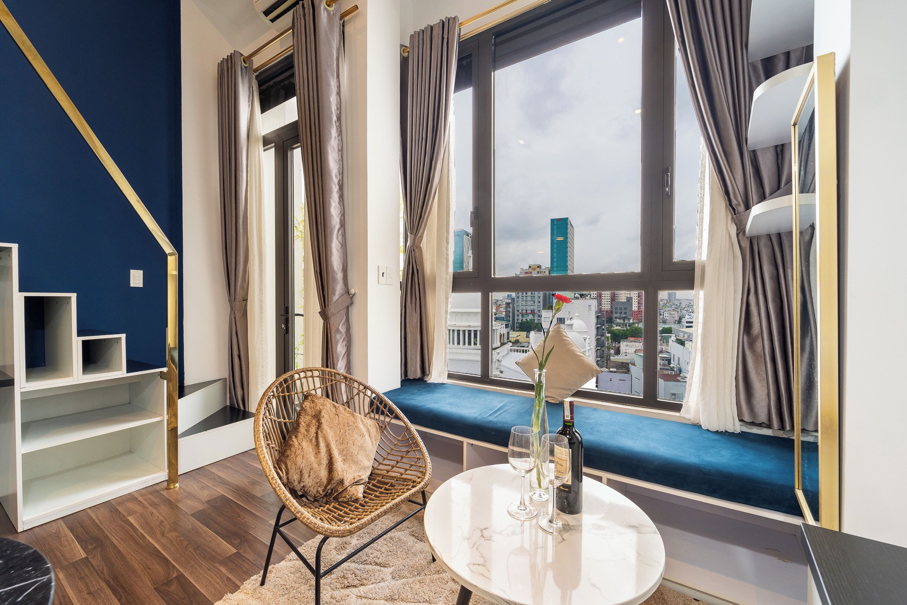Een kamer met een groot raam, een rieten stoel, een ronde tafel met een fles en glazen, en een blauwe bank.