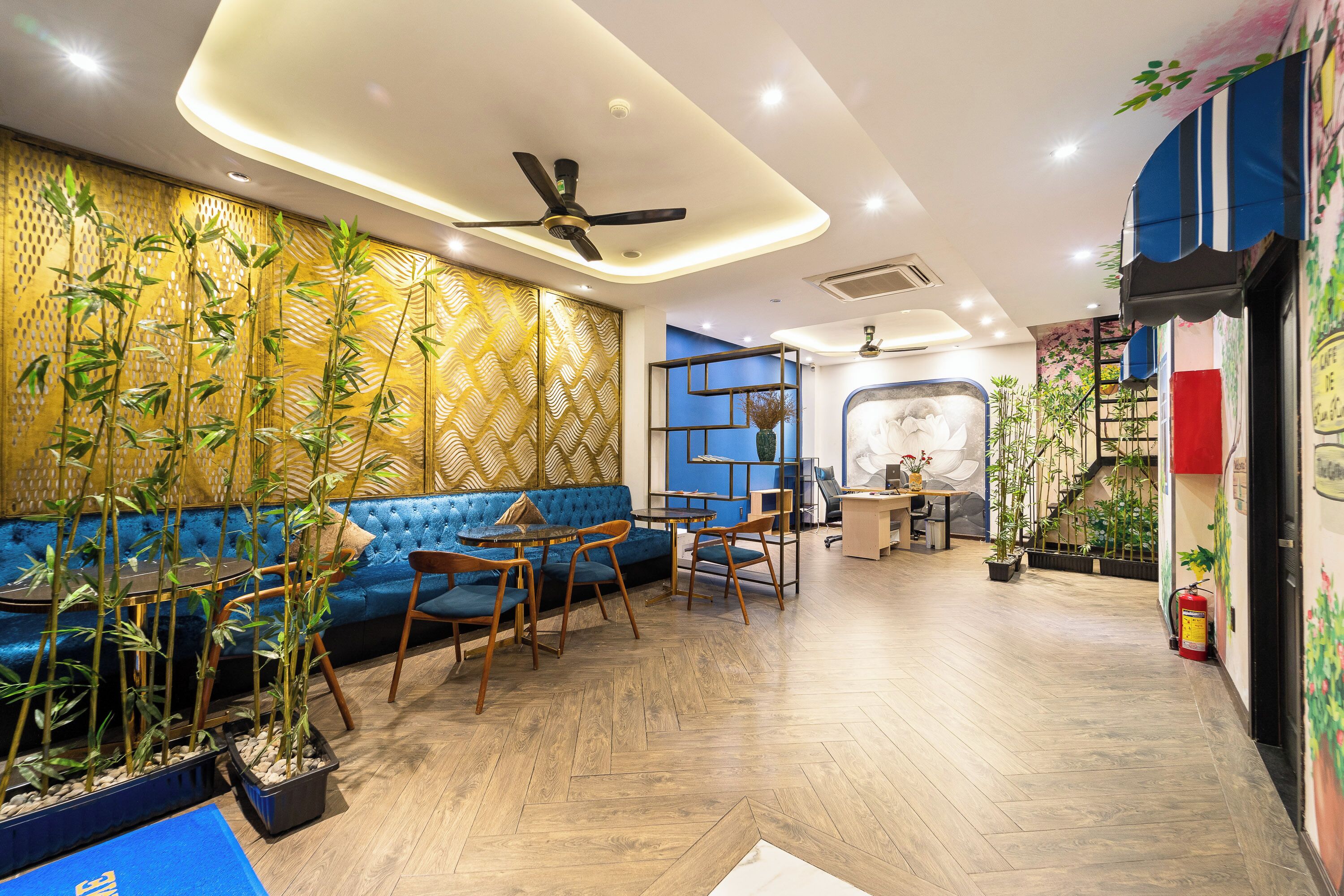 Un interior moderno con un sofá azul, sillas de madera, plantas de bambú y un ventilador de techo.