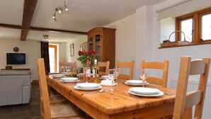 Un coin repas avec une table en bois dressée pour quatre personnes, entourée de chaises, d’un meuble et d’un téléviseur en arrière-plan.