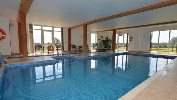Une piscine intérieure avec des parois en verre transparent, des poutres en bois et une vue sur l’extérieur.