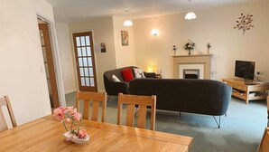 Een woonkamer met een bank, een open haard, een televisie en een eethoek met een tafel en stoelen.