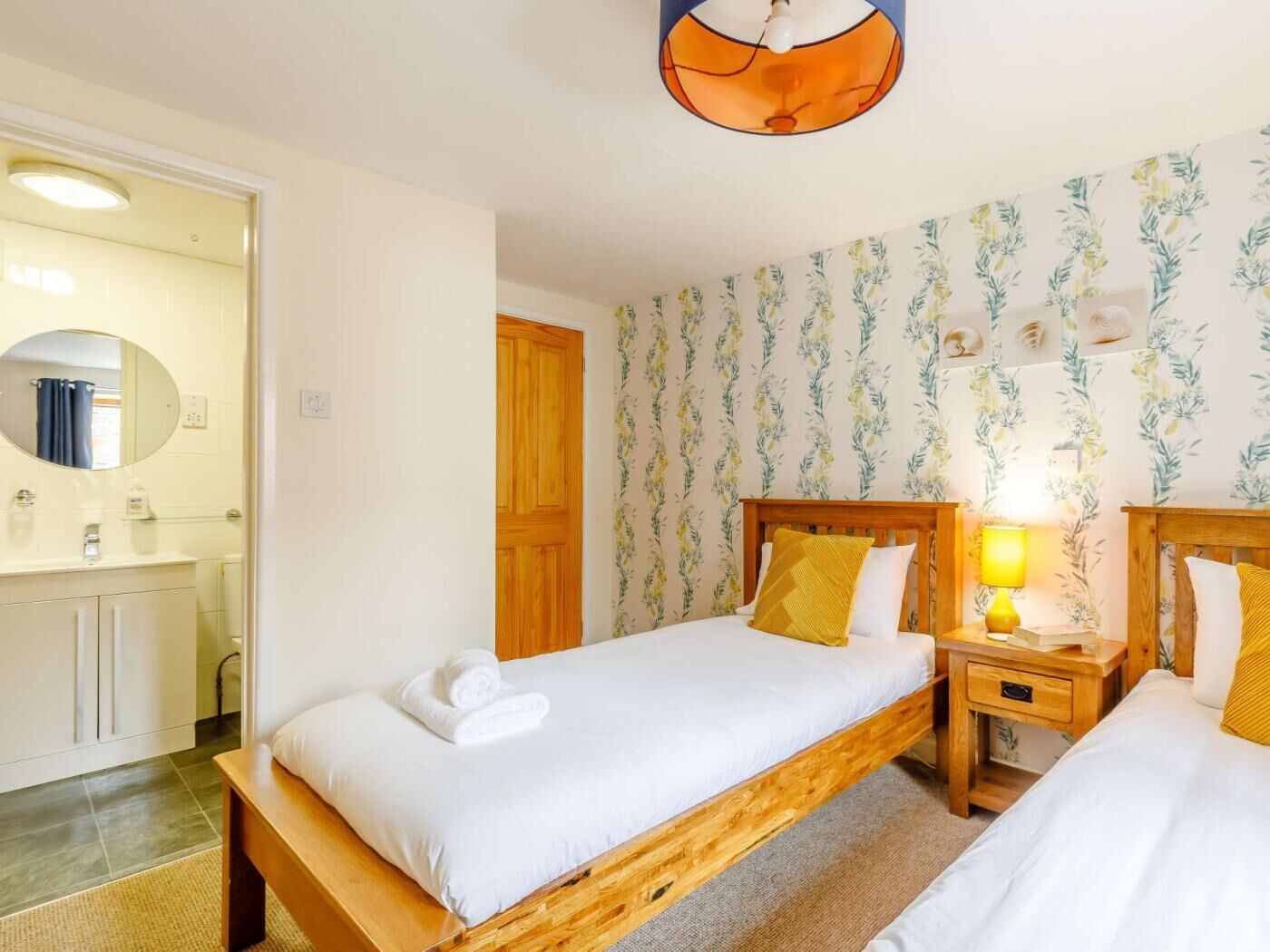 A bedroom with two beds, a wooden headboard, a nightstand with a lamp, and a bathroom visible through an open door.