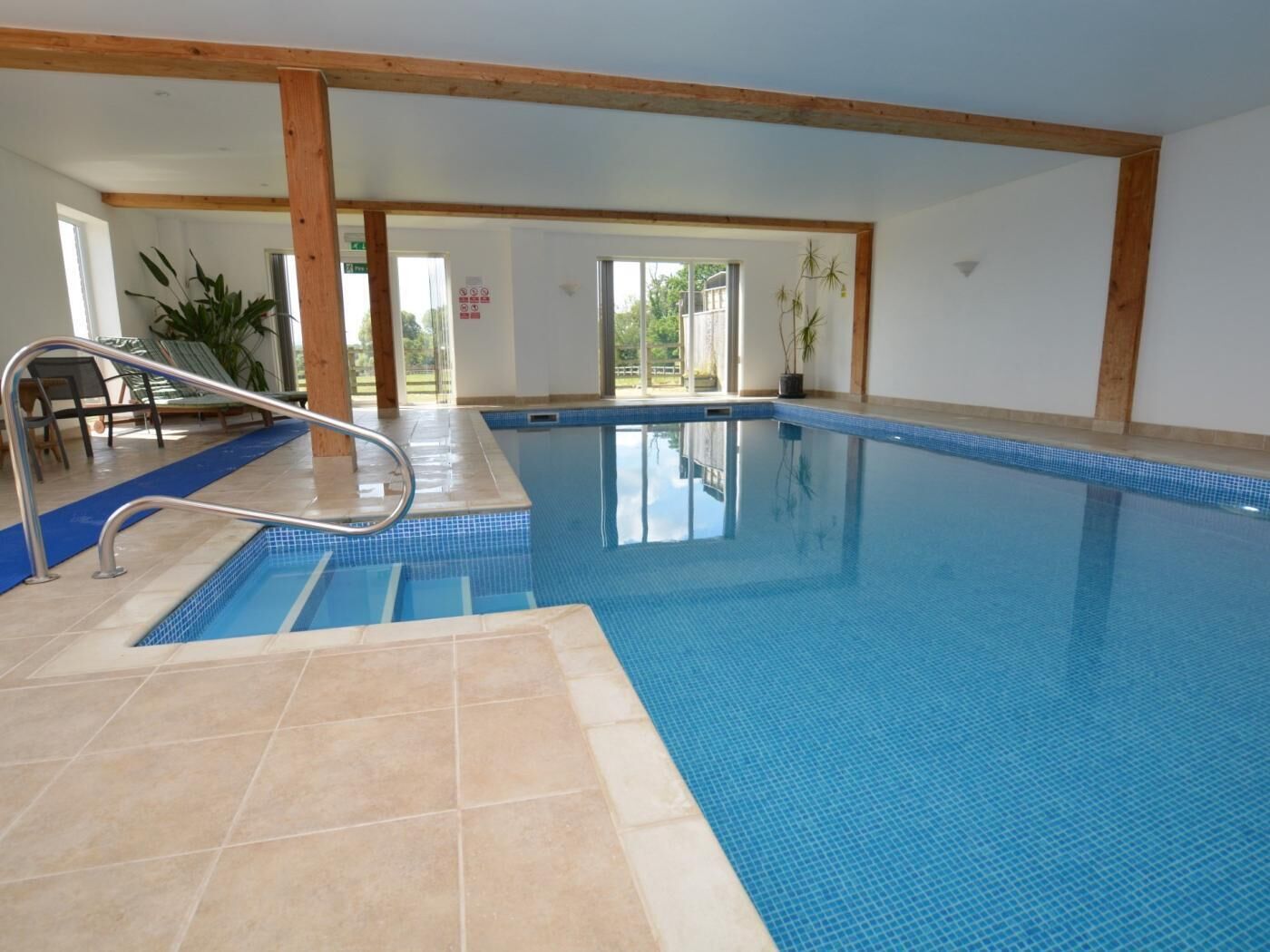 An indoor swimming pool with clear blue water, a metal ladder, and a view of the outside through large windows.