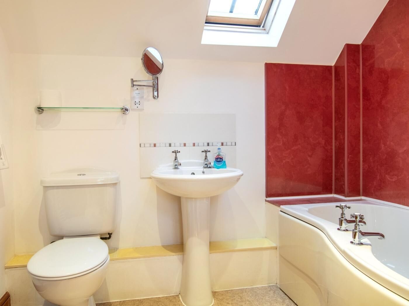 Une salle de bain comprenant des toilettes, un lavabo sur colonne, un miroir, une étagère en verre, un mur rouge et une baignoire.